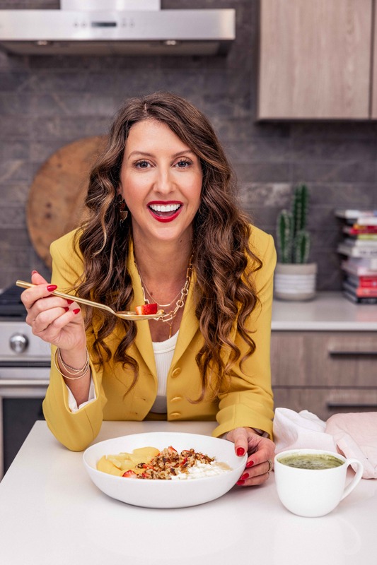 Simone Lovell in a bright yellow blazer leaning over a bowl, holding a gold spoon with a strawberry, a mug of matcha beside her